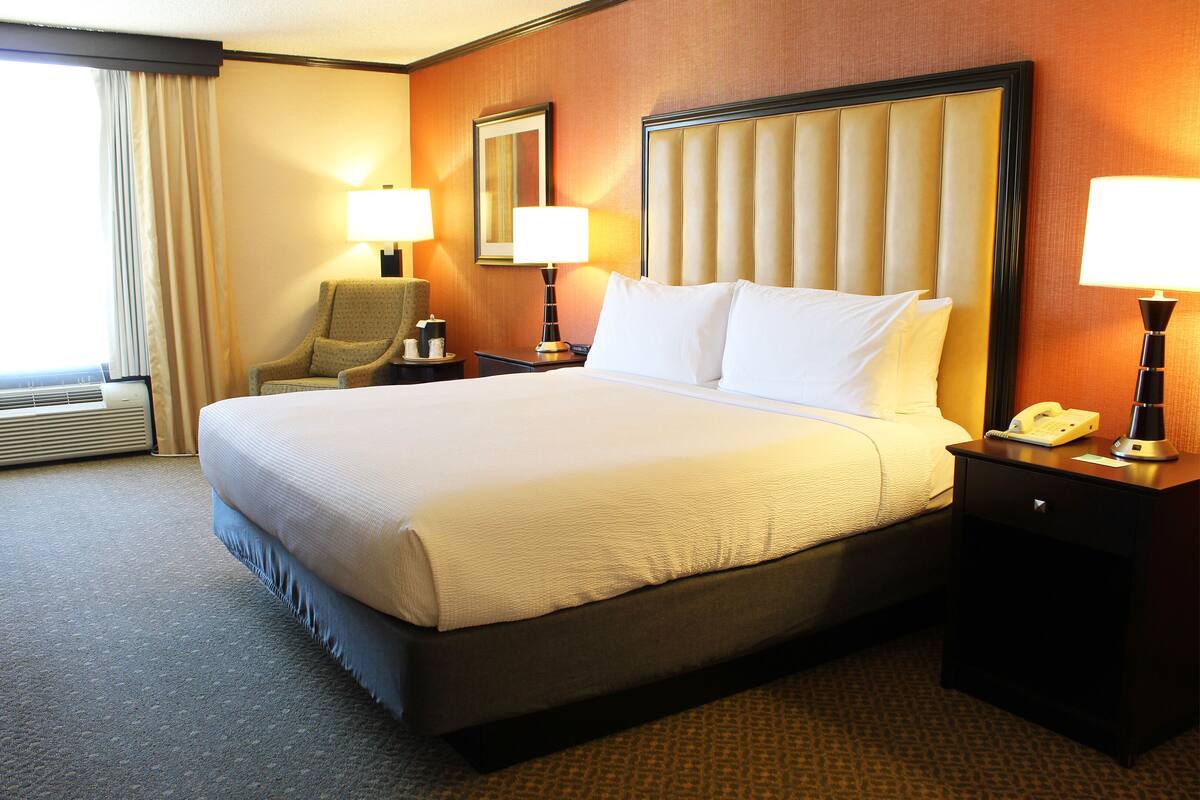 A hotel room with a large bed, two bedside lamps, a chair, a nightstand with a phone, and a window with curtains.