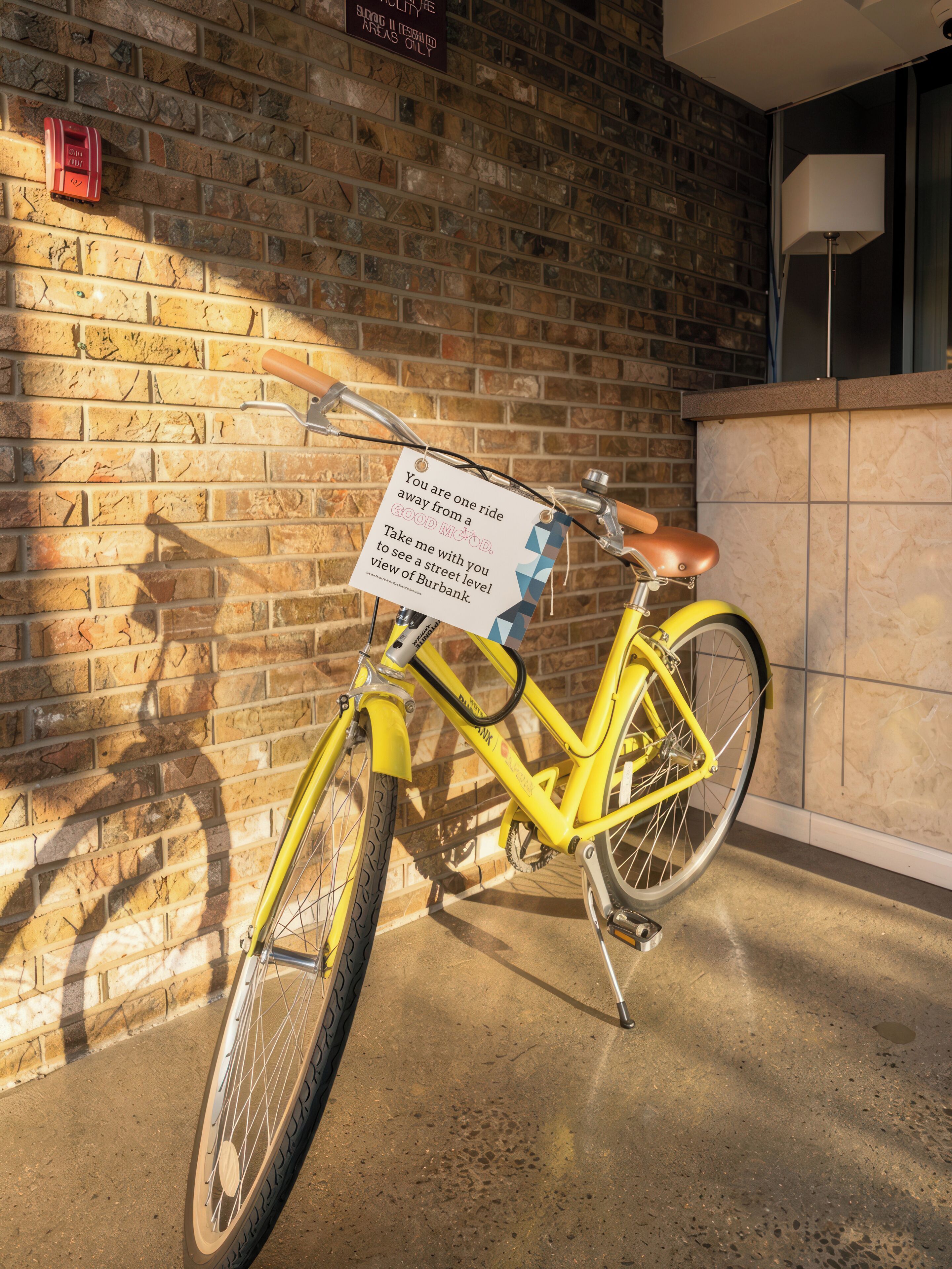 A yellow bicycle parked against a brick wall with a sign attached to the handlebars.