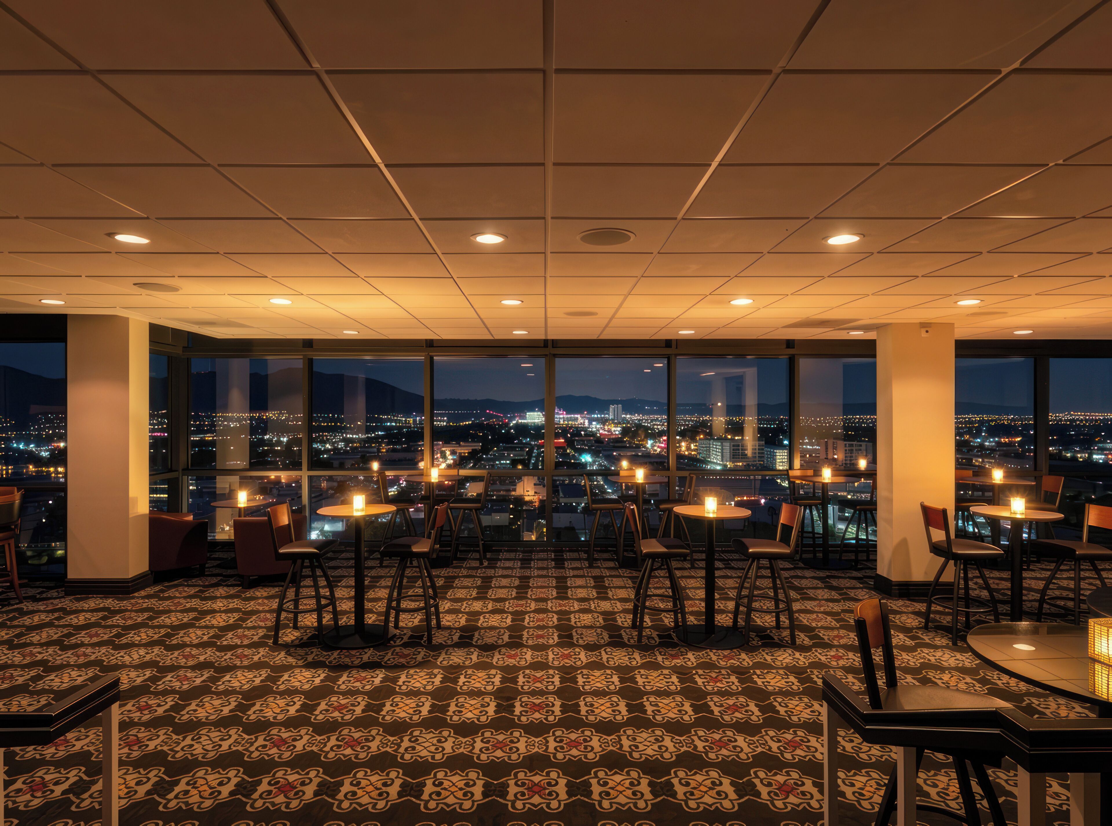 A high-rise hotel room with a panoramic city view, large windows, and a patterned carpet.