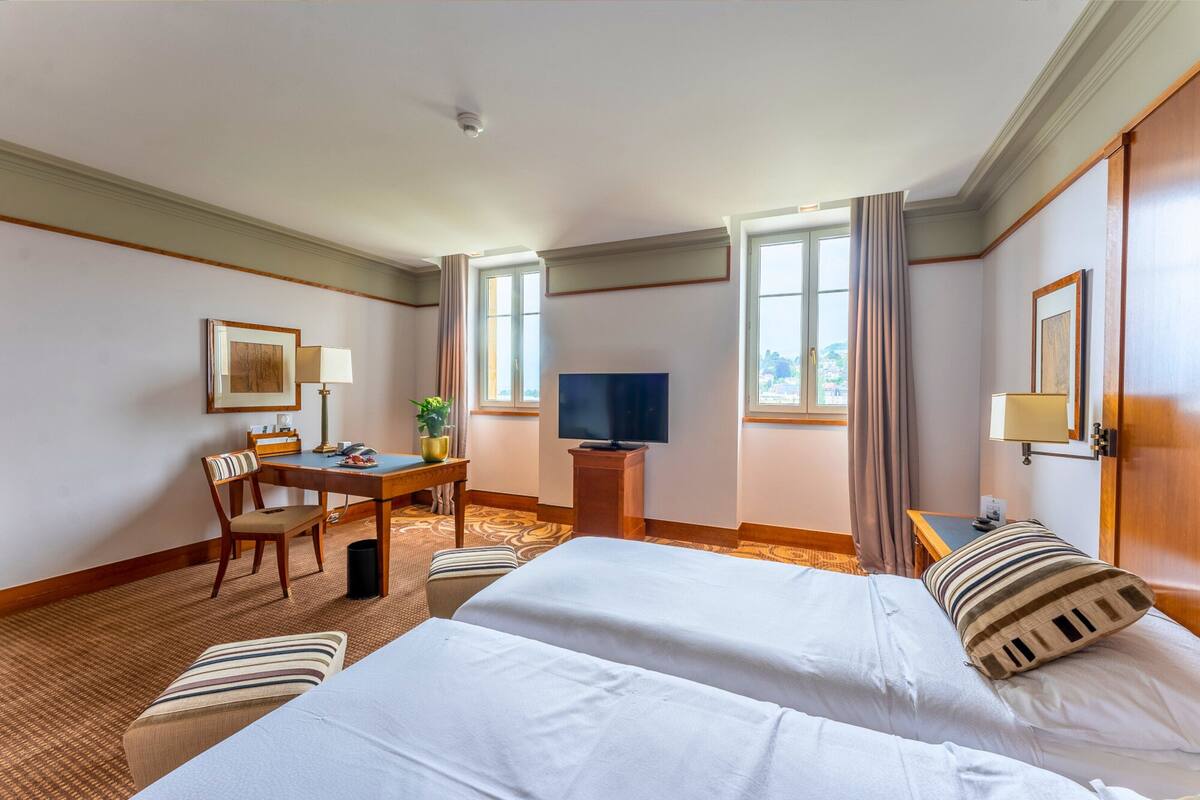 A hotel room with two beds, a desk with a TV, a chair, a small table, and a window with curtains.