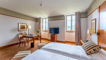 A hotel room with two beds, a desk with a TV, a chair, a small table, and a window with curtains.