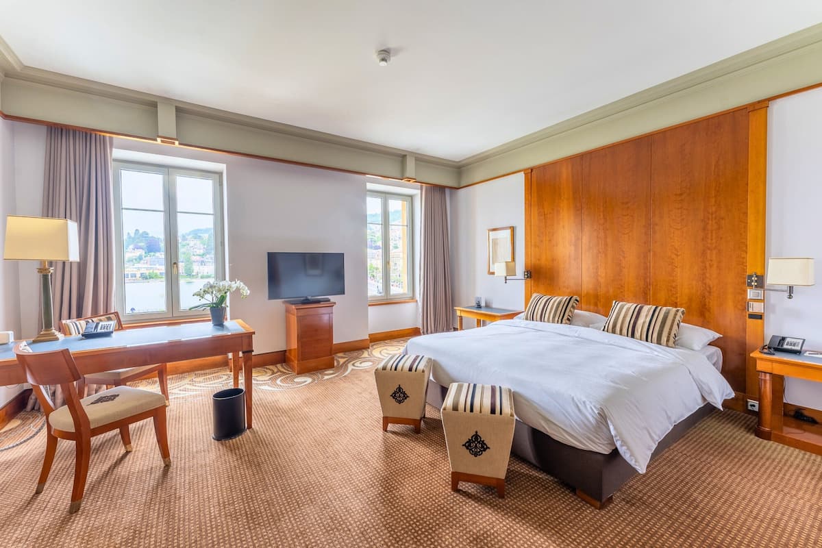 A hotel room with a large bed, a desk with a chair, a television, and a view of the city through the windows.