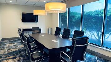 A conference room with a long table, black chairs, a TV on the wall, and large windows with a view of trees.