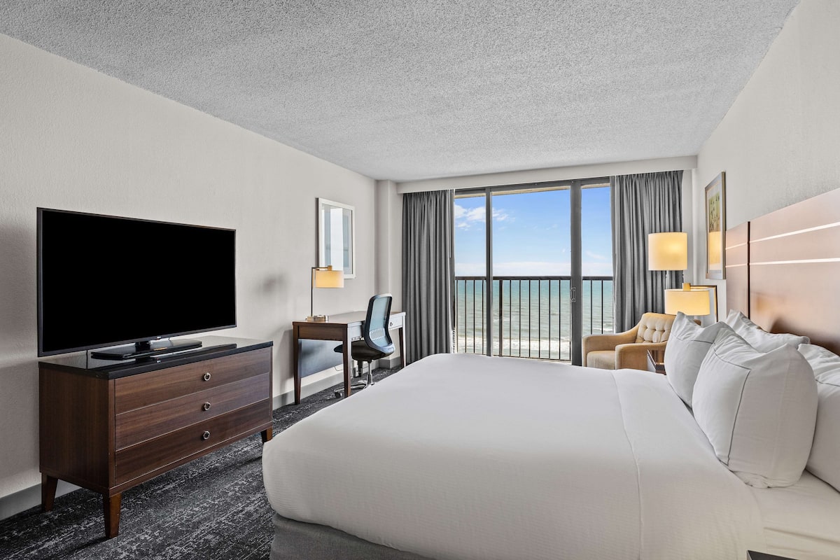 Habitación de hotel con una cama grande, un televisor de pantalla plana, un escritorio con una silla, una lámpara y vista al océano a través de la ventana.