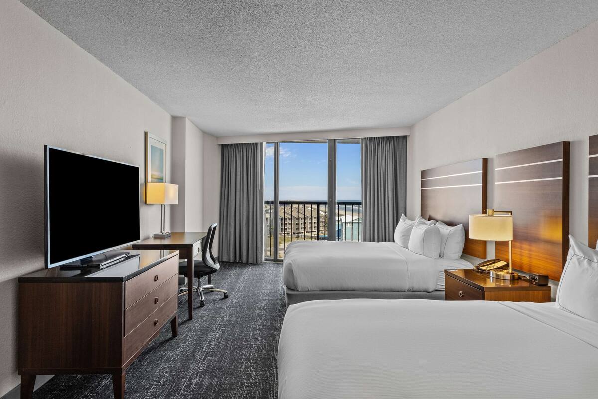 Habitación de hotel con una cama grande, una cama más pequeña, un escritorio con televisor, una lámpara y vistas a la ciudad.
