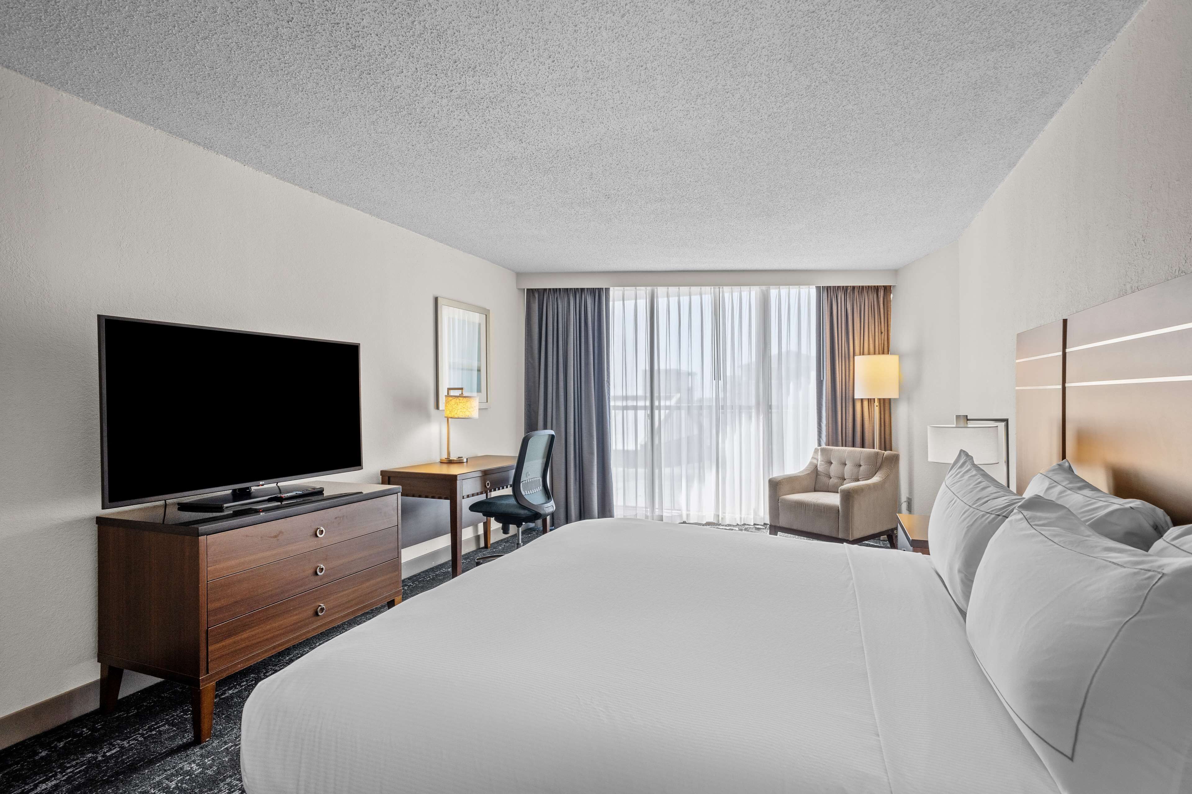 A hotel room with a large bed, a flat-screen TV, a desk with a chair, a lamp, and a window with curtains.
