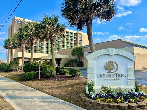 Entrada de un hotel con un cartel que dice "DOUBLETREE BY HILTON", rodeada de vegetación y palmeras.