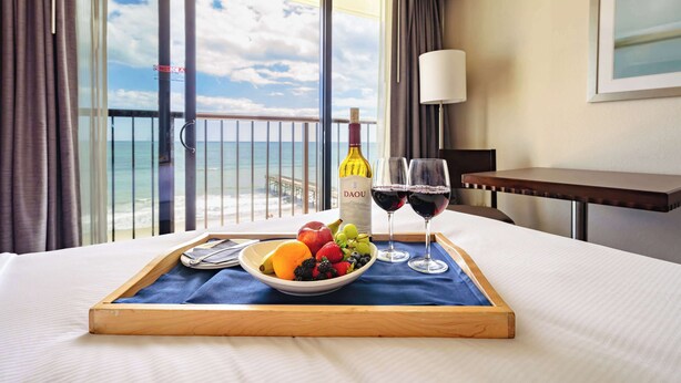Habitación de hotel con balcón, una cama, una bandeja con frutas y copas de vino, y una botella de vino.