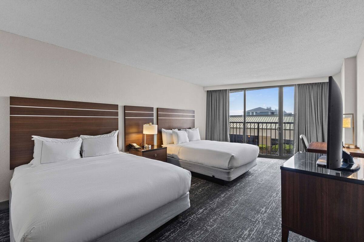 Habitación de hotel con dos camas, un escritorio, un televisor y un ventanal con vistas.