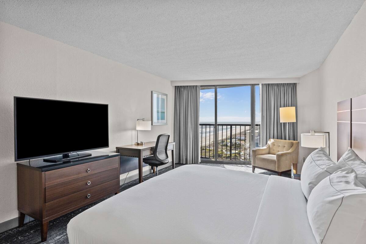Habitación de hotel con una cama grande, televisión, escritorio con silla y vista al océano a través de las ventanas.