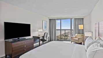 A hotel room with a large bed, a TV, a desk with a chair, and a view of the ocean through the windows.