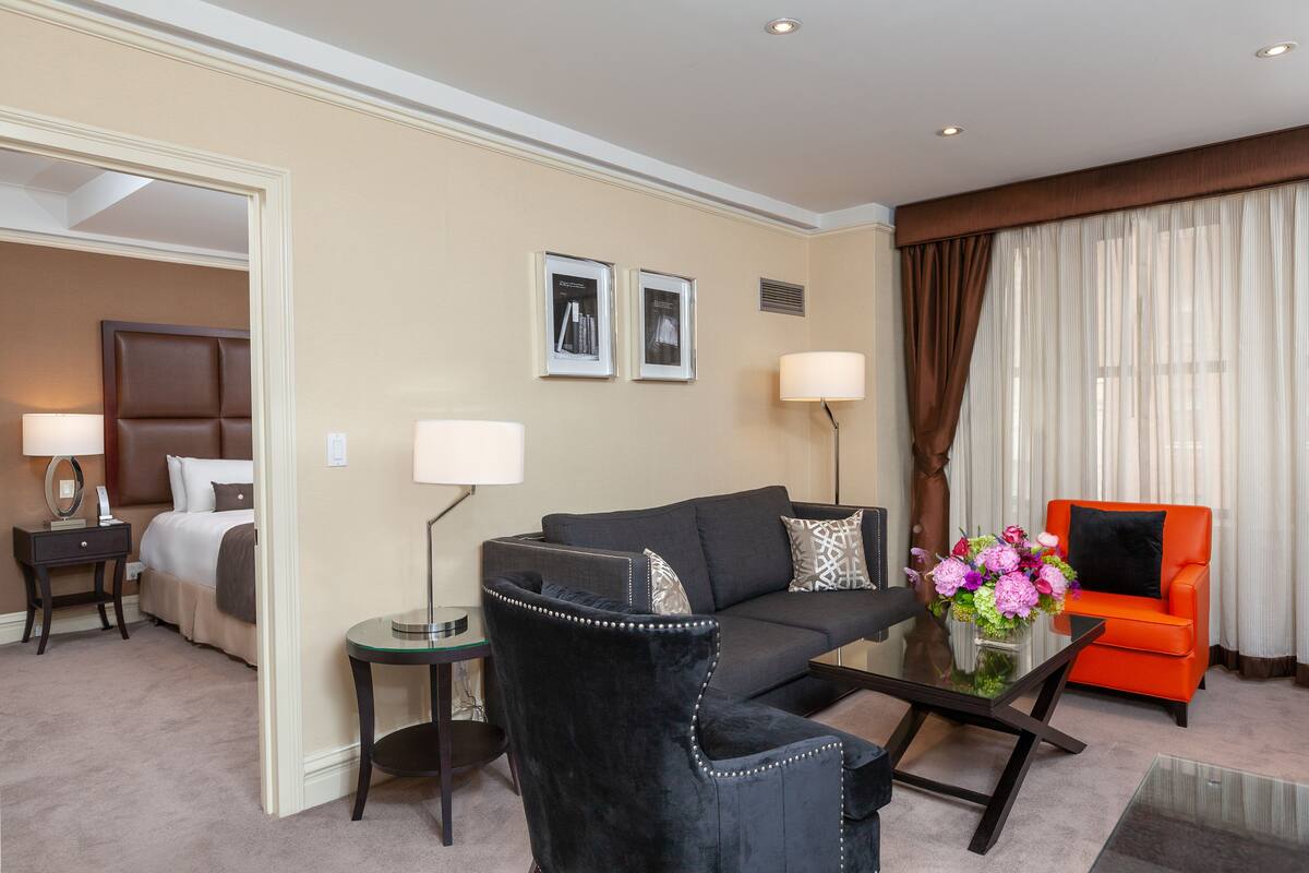 A hotel room with a bed, a black armchair, a sofa, an orange armchair, a glass coffee table, and two lamps.