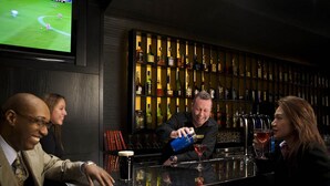 Zona de bar con una pantalla mostrando un evento deportivo, un barman preparando una bebida y una estantería con varias botellas.