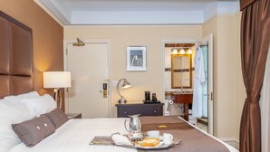 Una habitación de hotel con una cama bien hecha, una bandeja con artículos de desayuno, una cafetera y un baño visible a través de una puerta abierta.