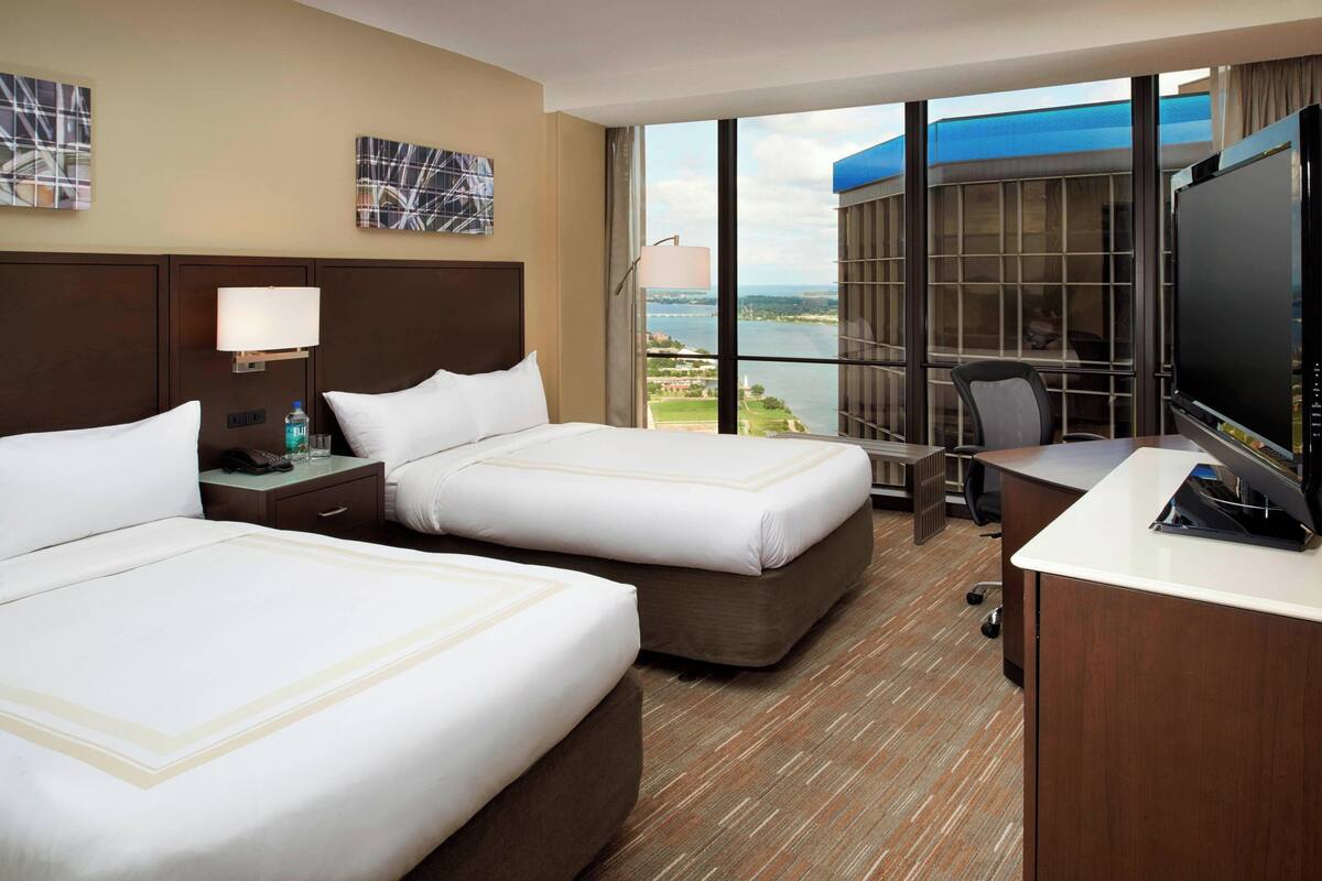 Habitación de hotel con dos camas, un escritorio, un televisor y un amplio ventanal con vista a un cuerpo de agua y edificios.
