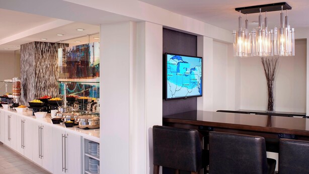 A modern bar area with a long counter, bar stools, a television displaying a map, and a decorative wall with vertical elements. - Detroit Marriott at the Renaissance Center (Detroit)