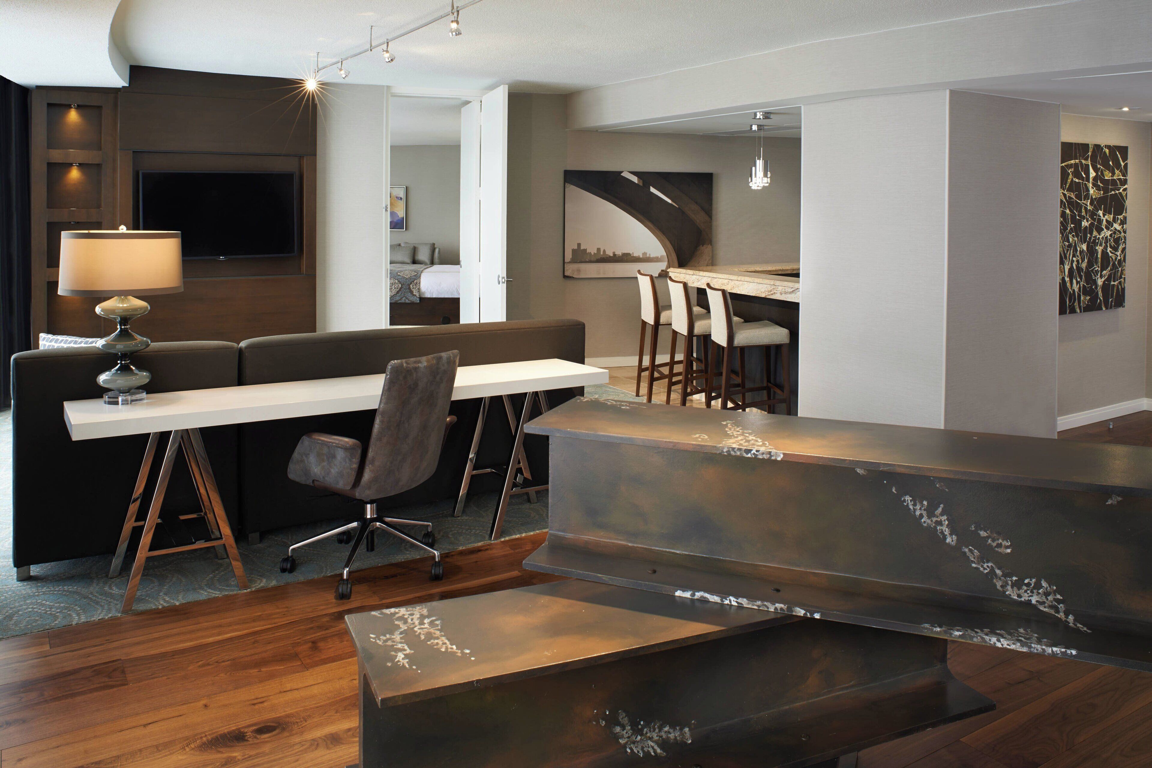 Una habitación de hotel moderna con suelo de madera, un escritorio blanco con lámpara, un sofá y una zona de bar con taburetes.