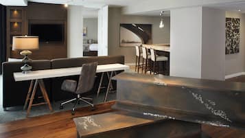 Una habitación de hotel moderna con suelo de madera, un escritorio blanco con lámpara, un sofá y una zona de bar con taburetes.