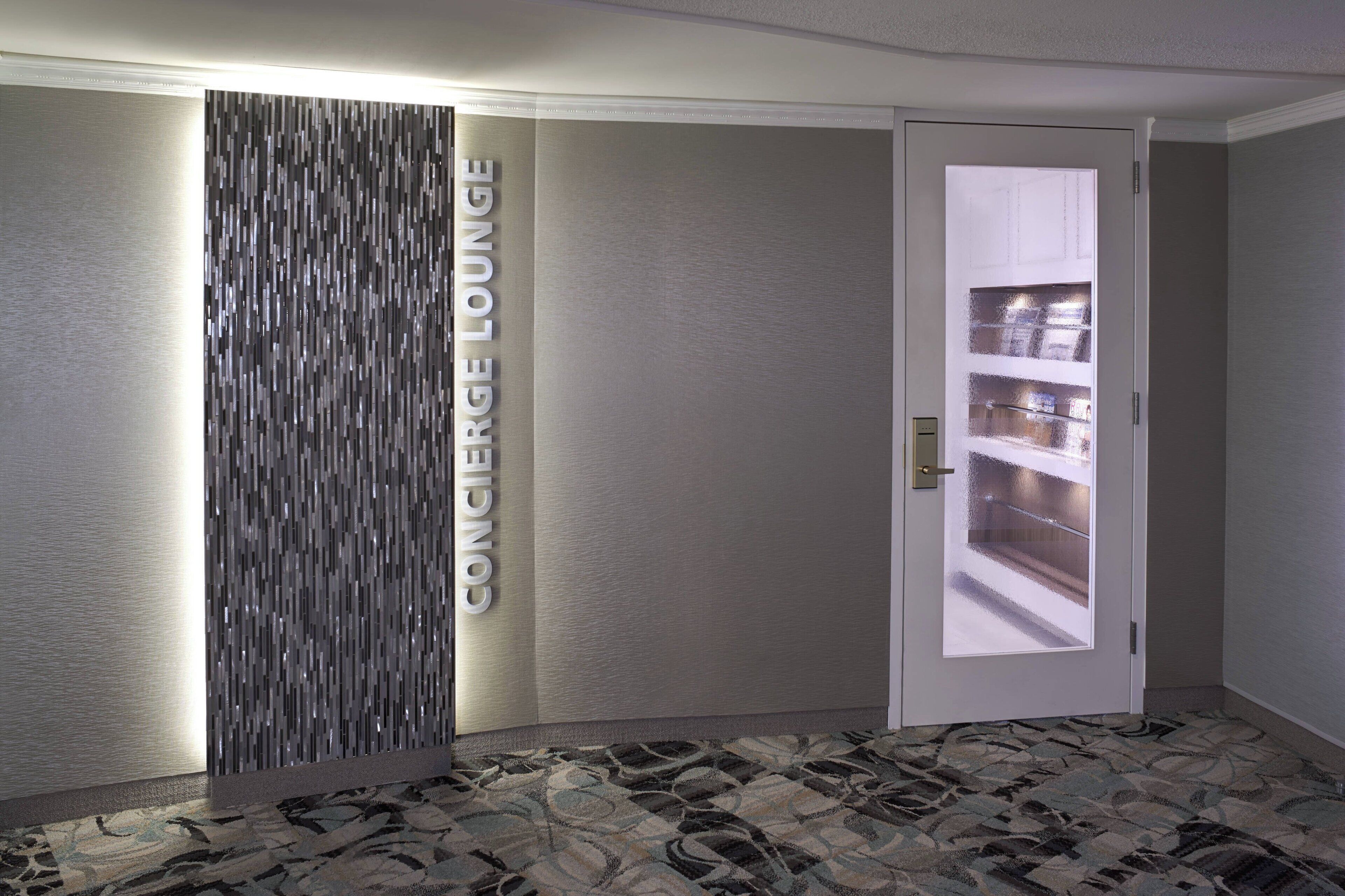 Interior moderno com um painel vertical com a inscrição 'CONCIERGE LOUNGE' e uma porta que dá para um espaço com prateleiras.