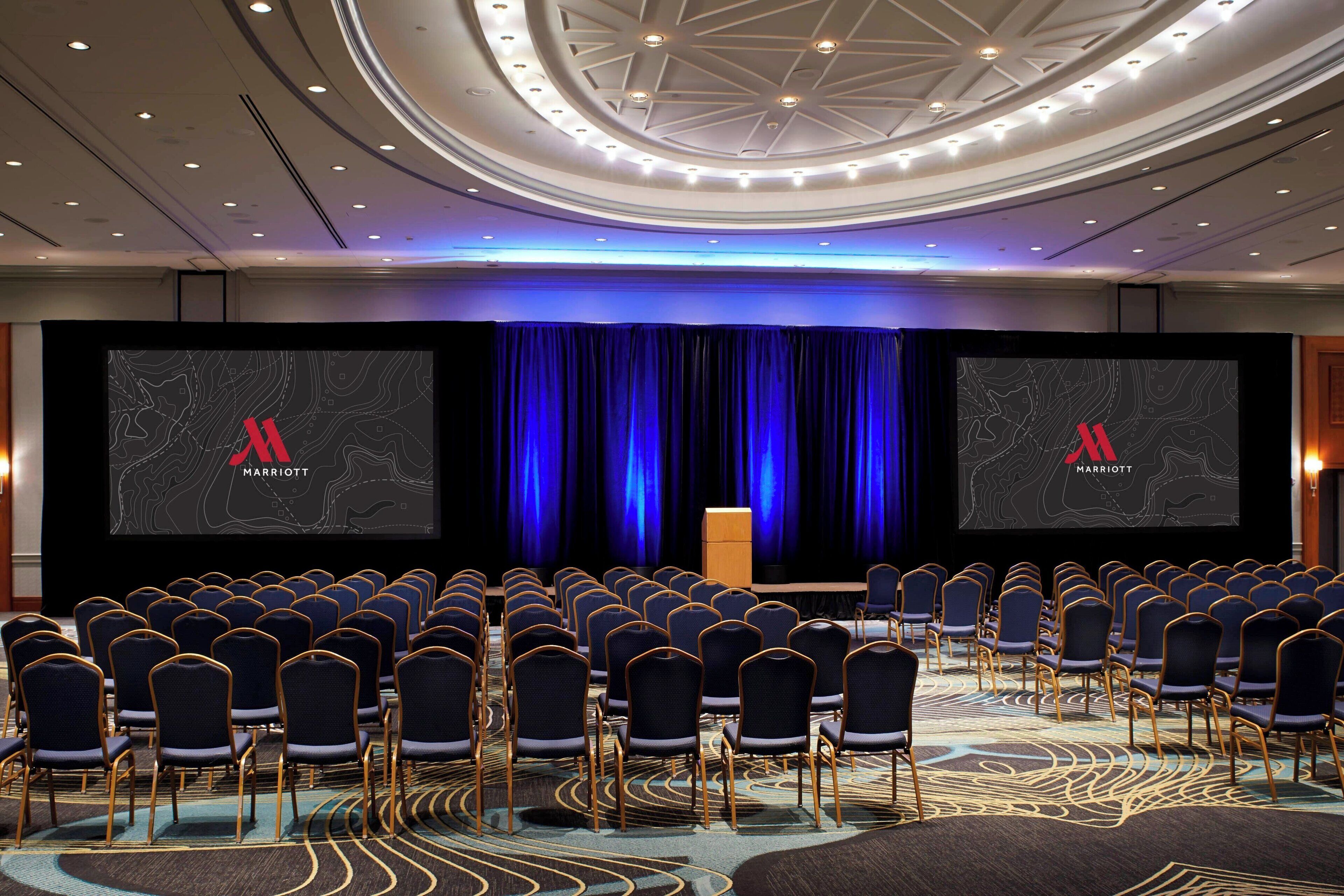 A conference room with rows of chairs, a stage with a blue curtain, and screens displaying the Marriott logo.
