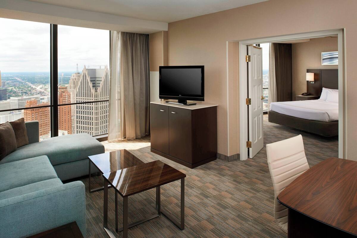 Habitación de hotel moderna con un ventanal amplio que ofrece vistas a la ciudad, un televisor de pantalla plana sobre un mueble de madera, un sofá, una mesita pequeña y una cama con una mesita de noche.