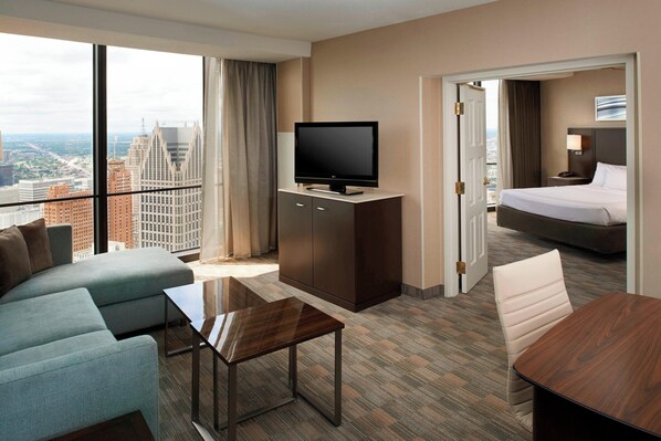 A modern hotel room with a large window offering a city view, a flat-screen TV on a wooden cabinet, a sofa, a small table, and a bed with a nightstand.