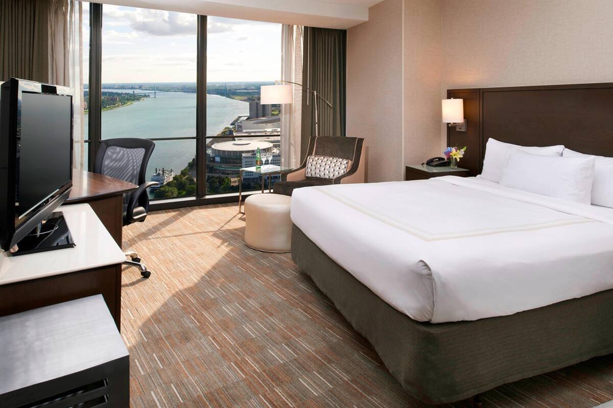 Habitación de hotel con una cama grande, un escritorio con televisor, una silla, una mesita de noche con lámpara, y vistas a un cuerpo de agua y edificios.