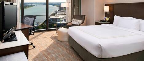 A hotel room with a large bed, a desk with a TV, a chair, a nightstand with a lamp, and a view of a body of water and buildings.