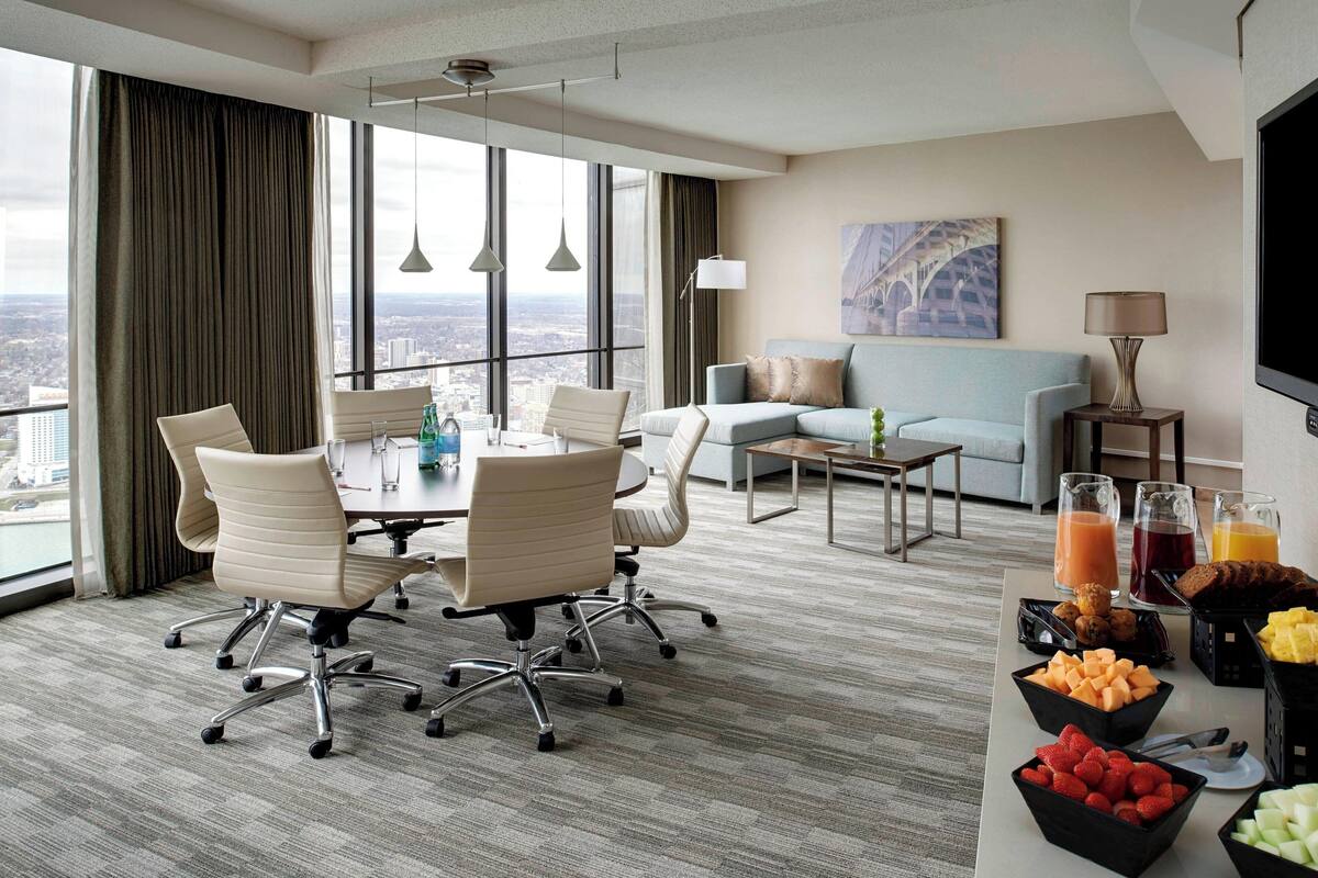 Una habitación de hotel moderna con una amplia mesa de comedor, asientos cómodos, un sofá, un televisor y vistas a la ciudad.