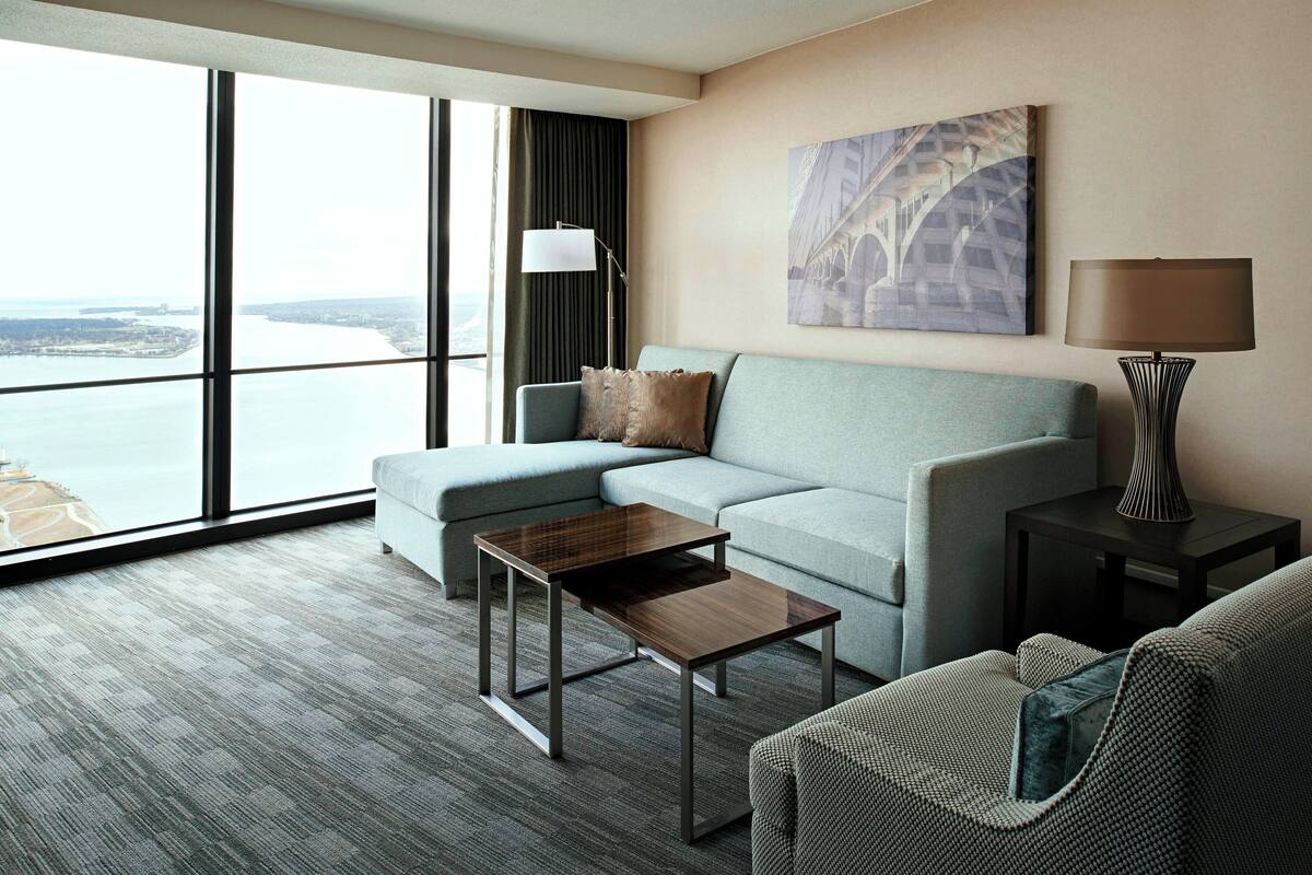 Habitación de hotel moderna con un amplio ventanal que ofrece vistas al agua y a un puente, un sofá gris, una pequeña mesa de madera y una lámpara en una mesita auxiliar.