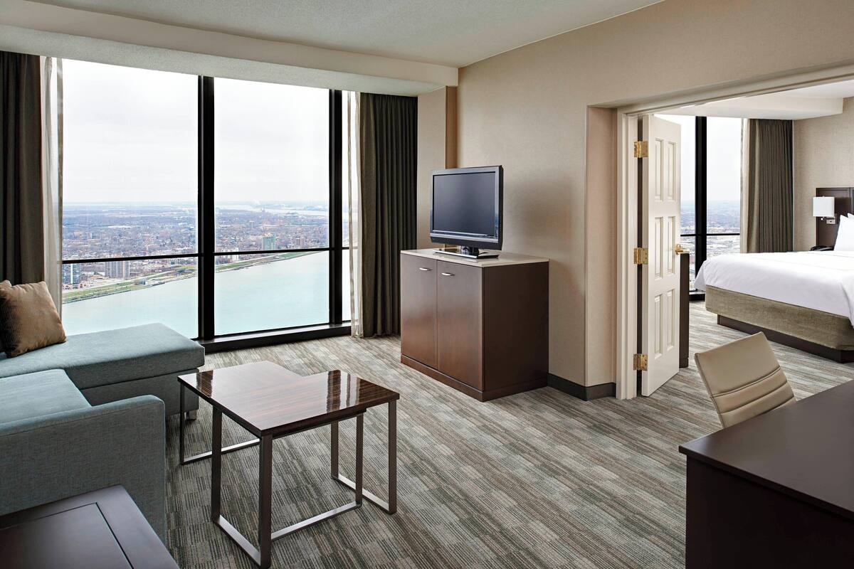 Una habitación de hotel con un ventanal grande, un televisor de pantalla plana, una cama, un sofá, una mesa de centro y un escritorio.
