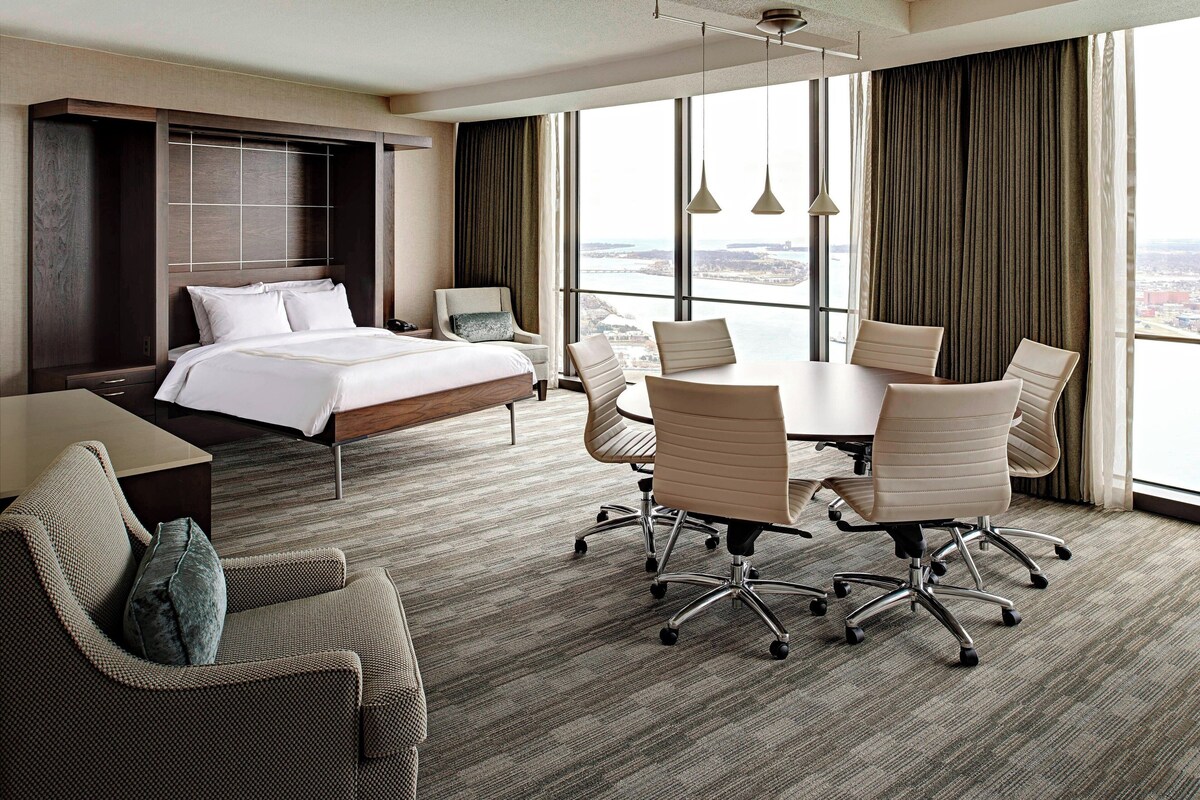 A hotel room with a large bed, a dining table with chairs, a sofa, and a view of the city through large windows.