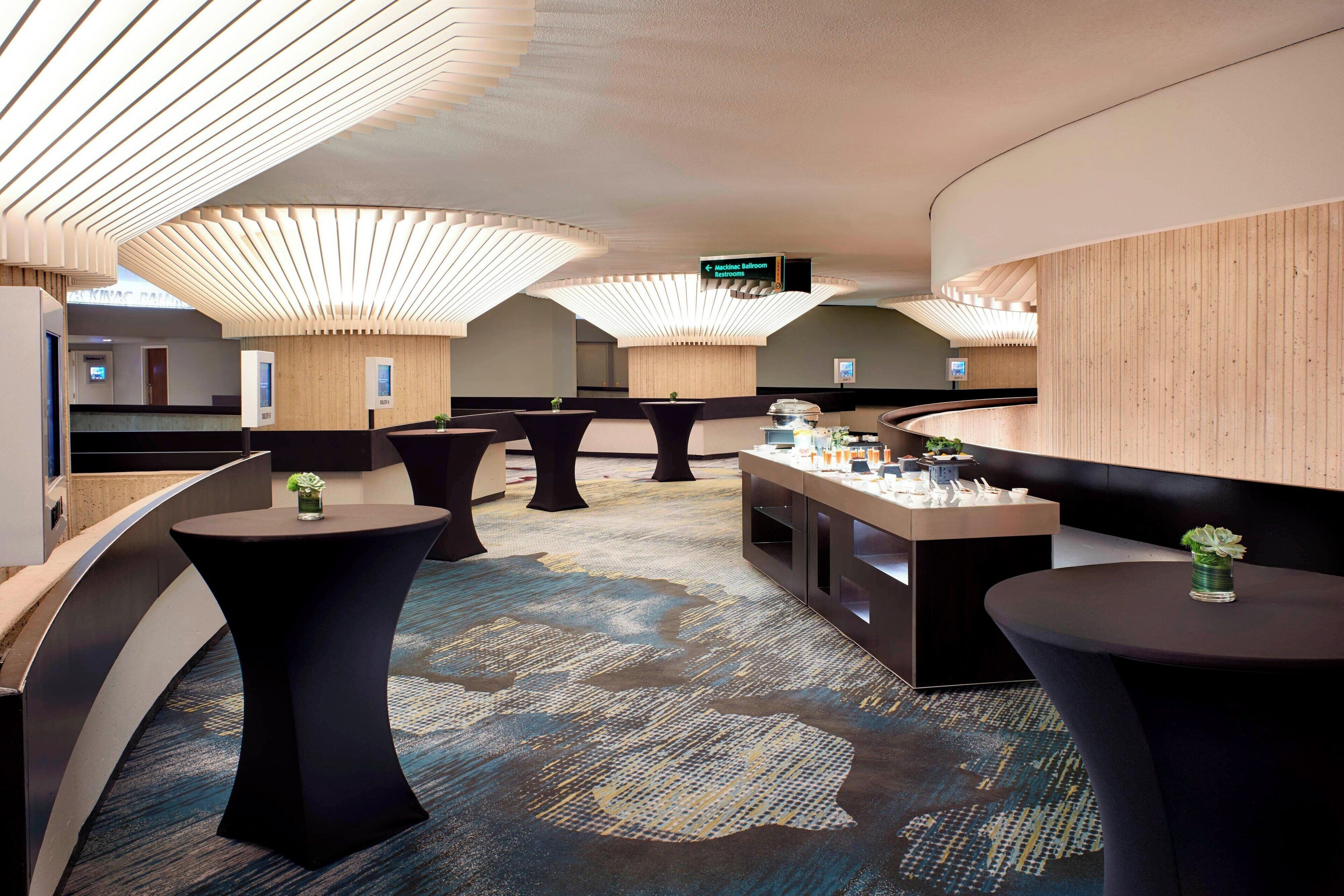 A modern hotel lobby with round tables, a reception desk, and a sign indicating the direction to the bathrooms.