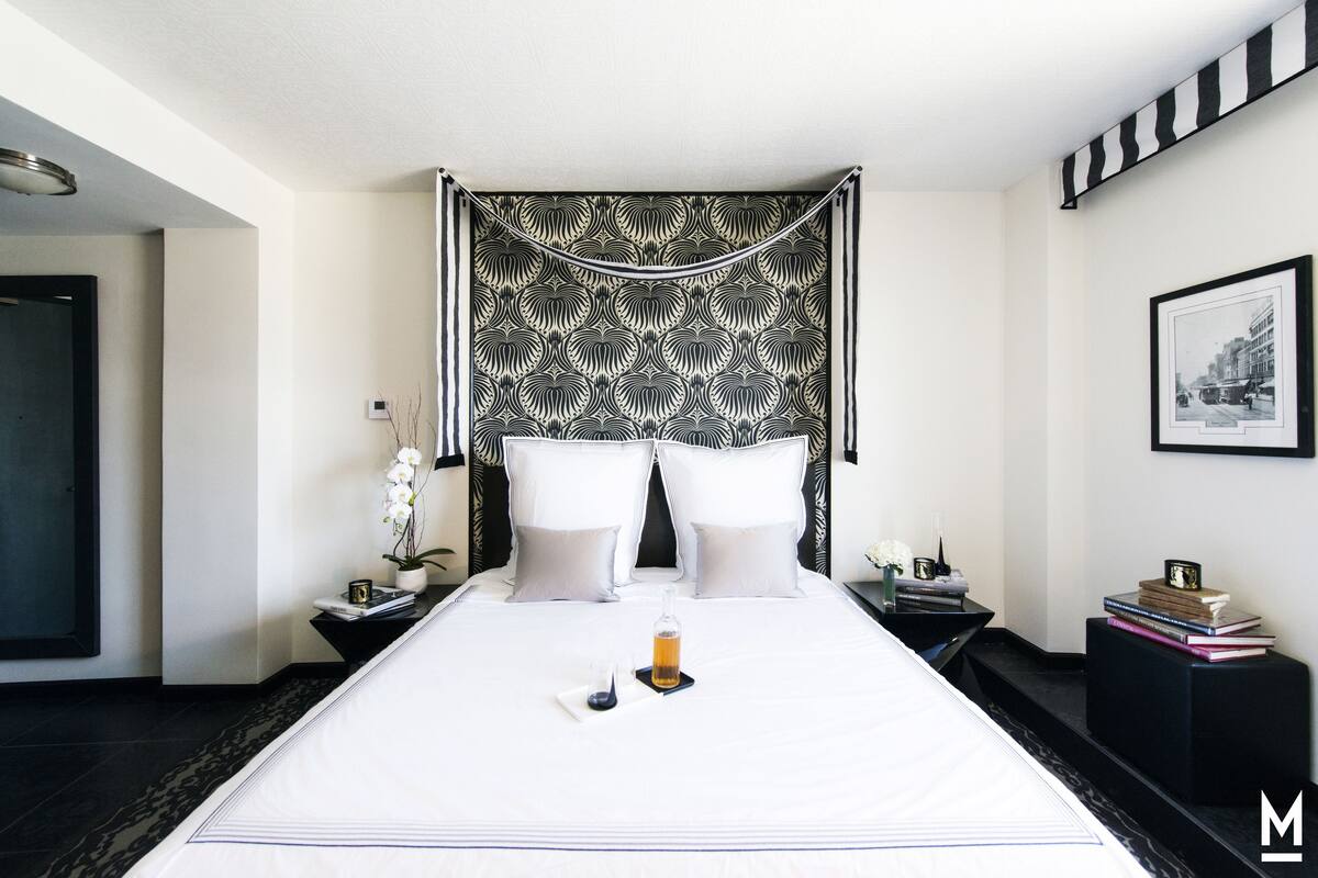 Una cama bien hecha con ropa blanca, una mesita de noche con una lámpara y un cuadro en la pared.