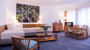 Quarto de hotel com sofá, mesa de centro de madeira, mesa de jantar com cadeiras, televisão e uma pintura grande na parede.