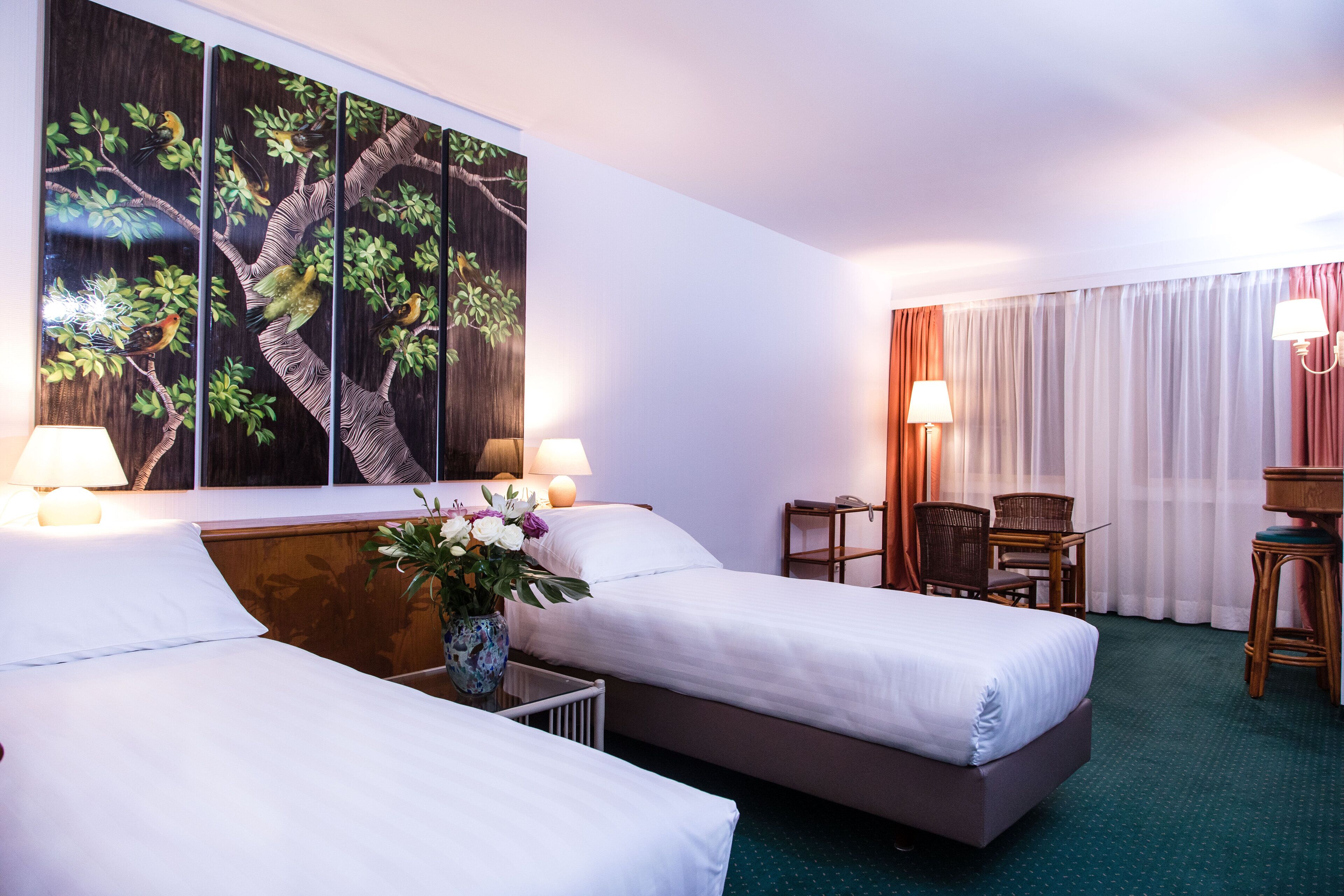 A hotel room with two beds, a desk, a chair, a lamp, and a large painting of birds on a tree.
