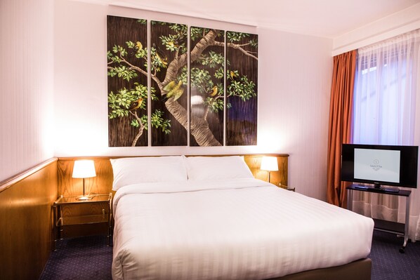 Une chambre d’hôtel avec un grand lit, deux lampes de chevet, un téléviseur à écran plat et une œuvre d’art fixée au mur représentant un arbre.