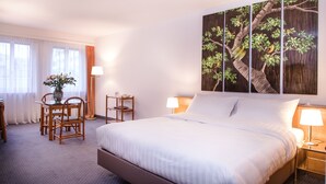 Quarto de hotel com uma cama grande, área de jantar com cadeiras e uma mesa, e um quadro de uma árvore pendurado na parede.