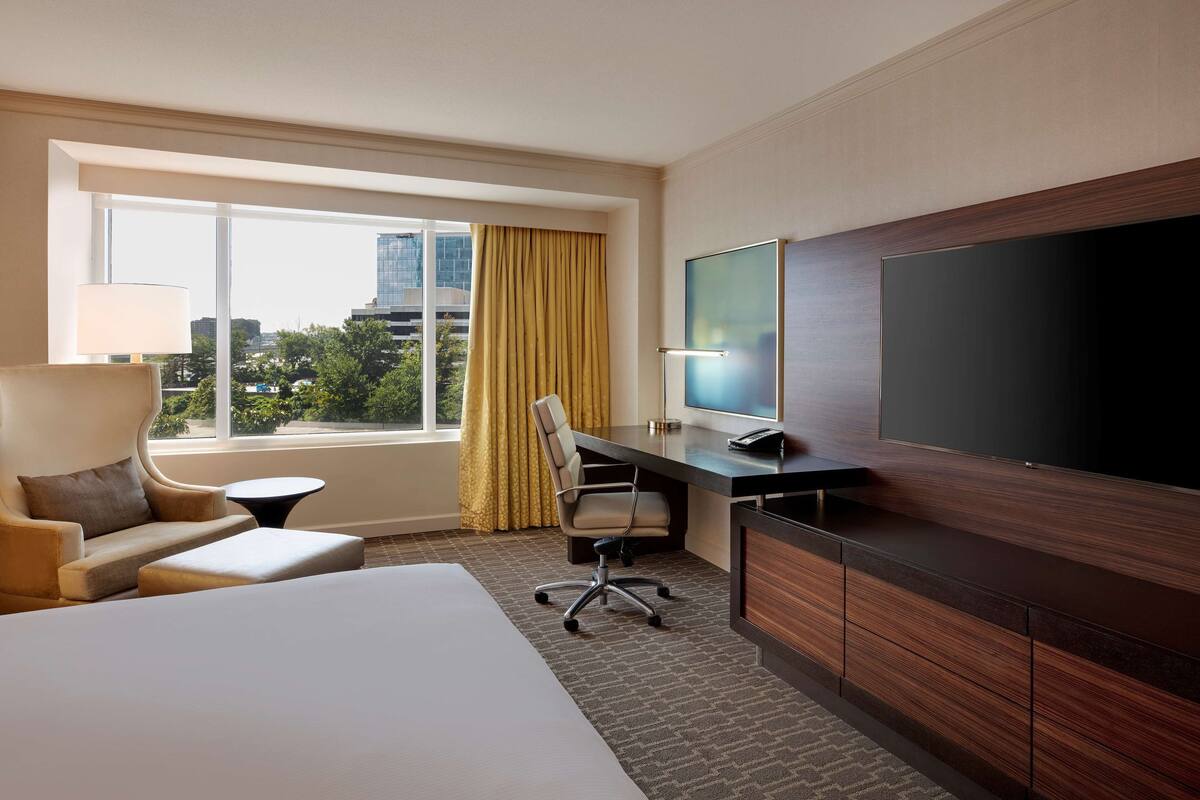 A hotel room with a large bed, a desk with a computer, a TV, and a view of the city through a window.