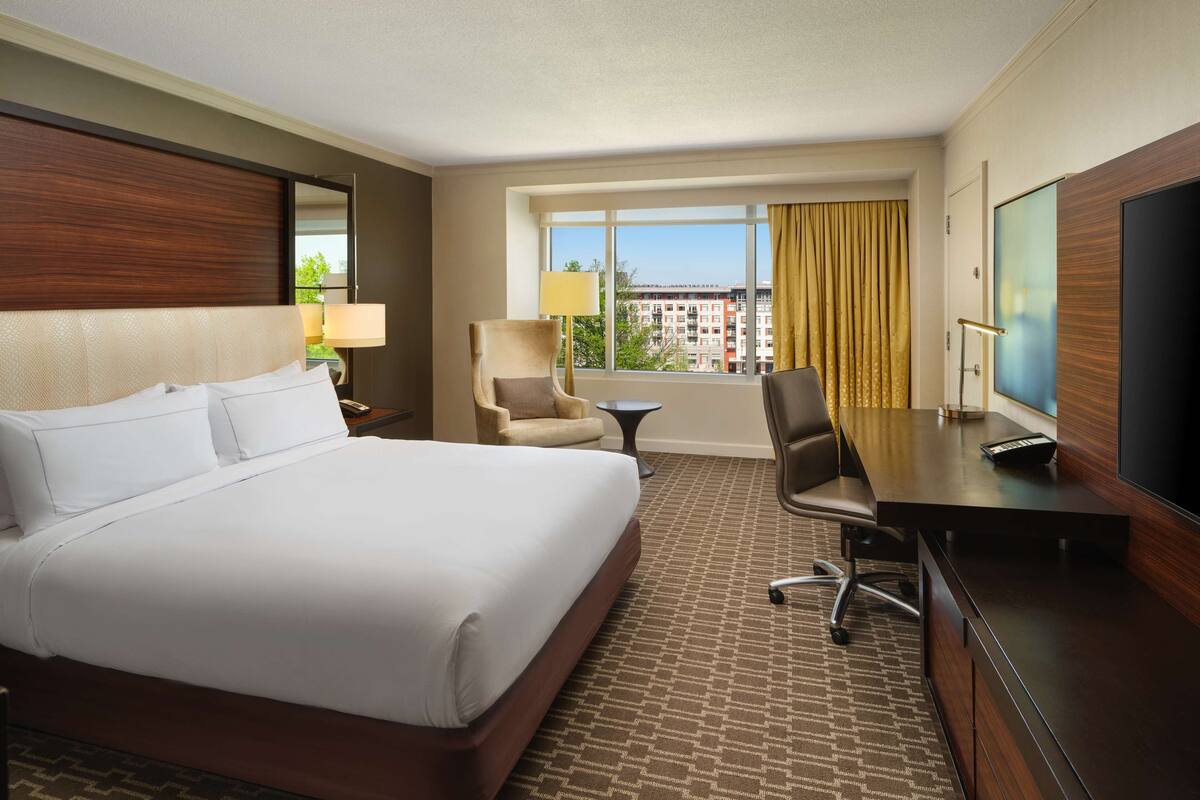 A hotel room with a large bed, a desk, a chair, a TV, and a window with a view of buildings.