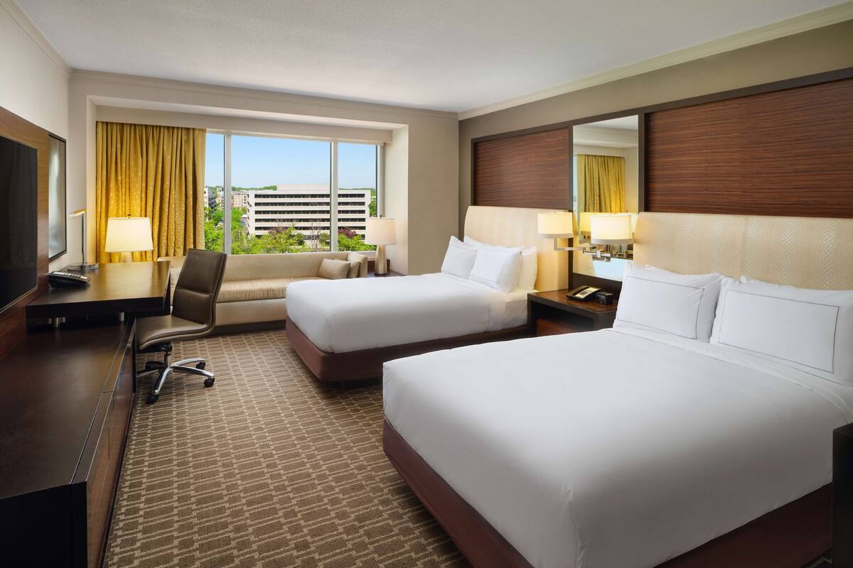 A hotel room with two beds, a desk, a chair, a TV, and a large window with curtains.