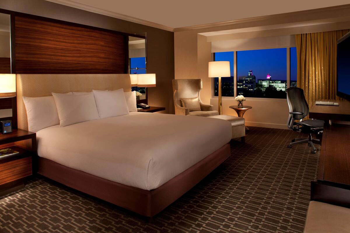 A hotel room with a large bed, a desk, a chair, and a view of the city at night.