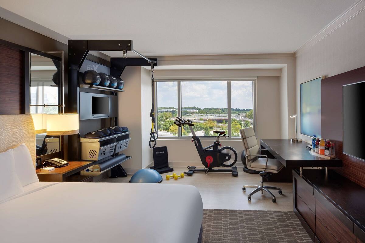 A hotel room with a bed, desk, TV, exercise equipment, and a large window with a city view.