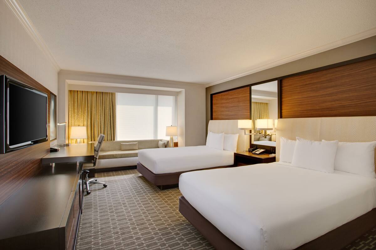 A hotel room with two beds, a desk with a flat-screen TV, a sofa, and a large window with curtains.