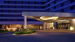 Una entrada de hotel moderna con un elemento de iluminación circular, un jardín ajardinado y un edificio de varios pisos con techo plano.