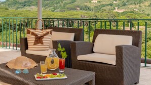 Uma varanda com móveis de vime, uma mesa com bebida e frutas, e vista para uma colina com edifícios.