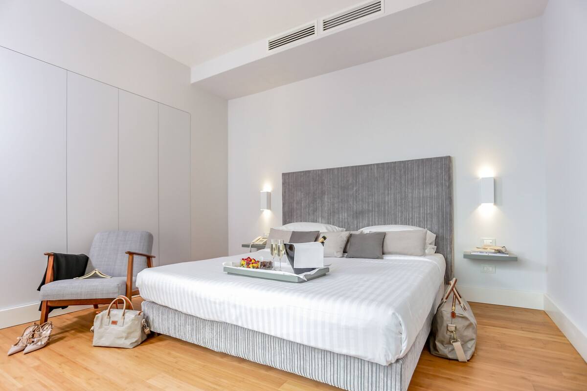 A modern hotel room with a large bed, a chair, a bedside table with a lamp, and a tray with toiletries.