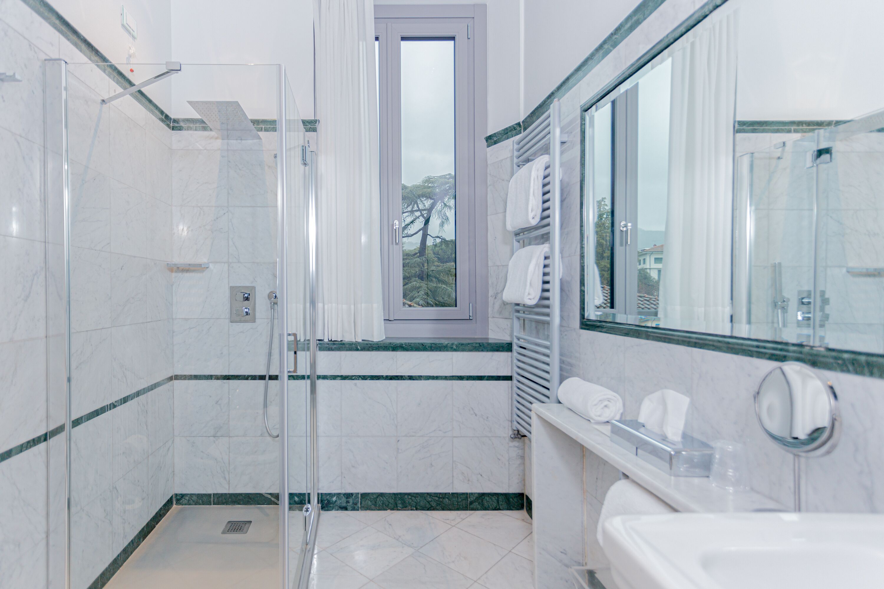 Une salle de bain moderne avec une douche à l’italienne en verre, un grand miroir et une fenêtre donnant sur la verdure.