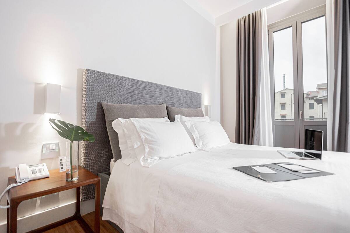 A hotel room with a bed, bedside table, lamp, and a view of buildings through the window.