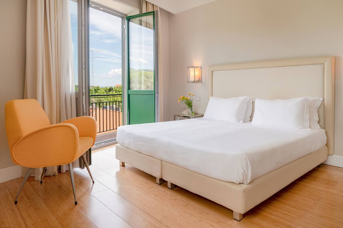 A hotel room with a large bed, an orange chair, a small table with a vase, and a balcony with a view.